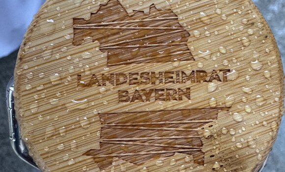 Zu sehen ist das LHR Logo eingraviert in einen Holzdeckel einer Trinkflasche mit Regentropfen drauf. 